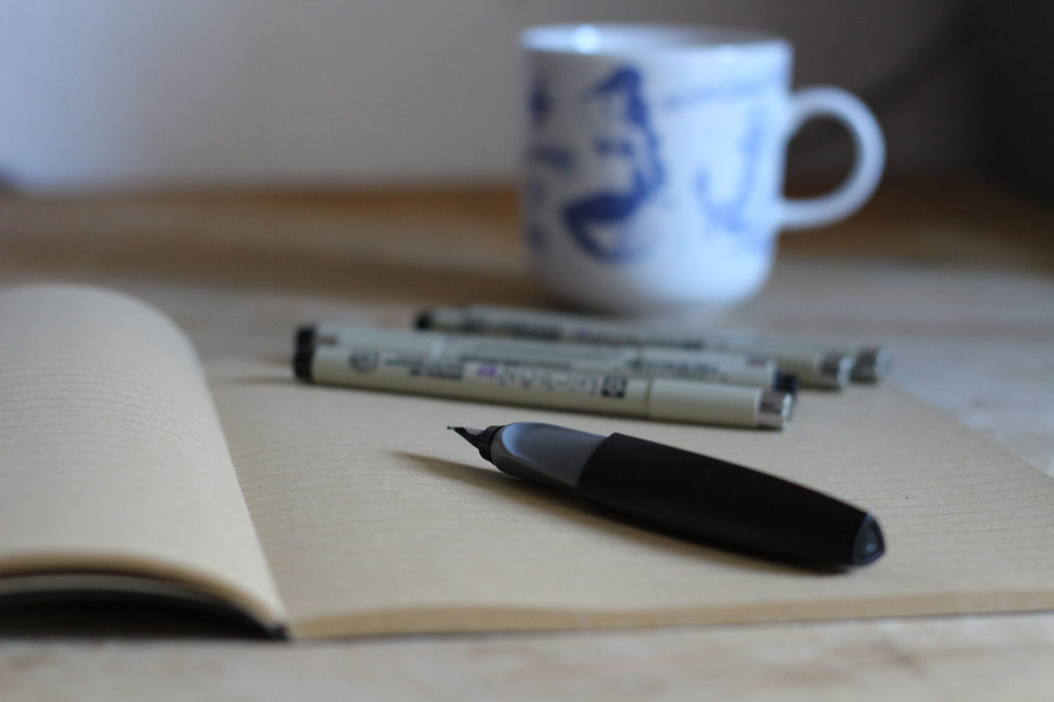 Das Bild zeigt eine Impression: Mein Schreibtisch mit einem Notizheft, einem Füller, Finelinern und einer Kaffeestasse im Hintergrund.
