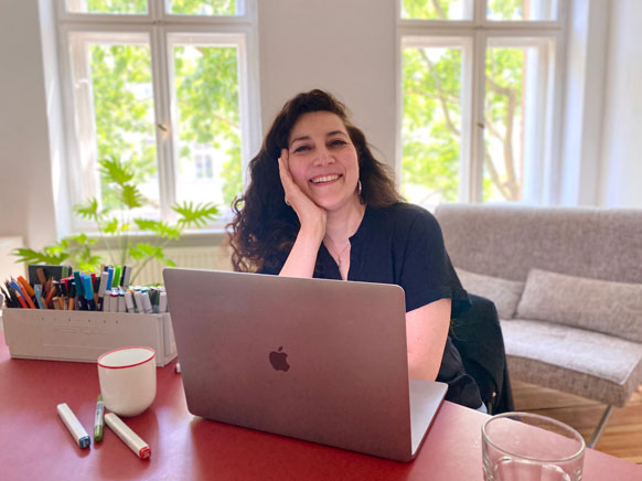 Auf dem Bild sieht man die Texterin Samira Suweidan in ihrem Büro in Berlin. Sie sitzt am Schreibtisch vor dem Laptop und lächelt in die Kamera.